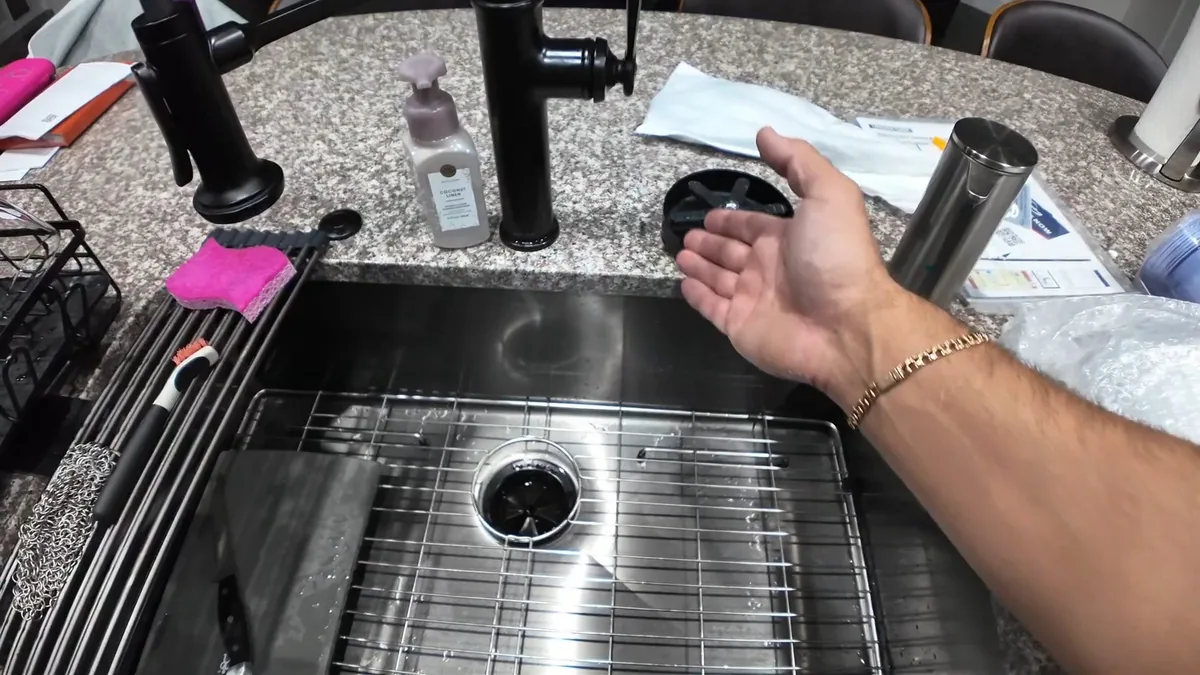Arm resting at the edge of a stainless kitchen sink with a black faucet and drain strainer