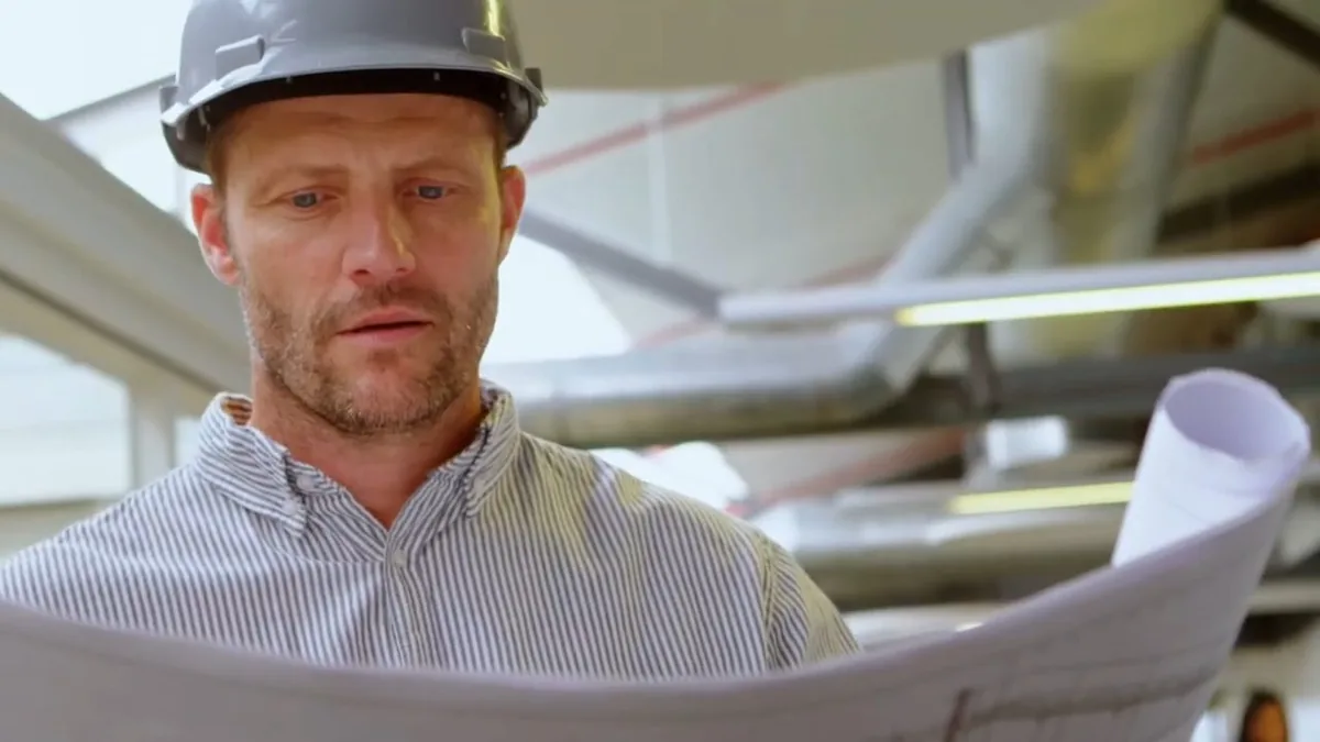 Man in a hard hat reading rolled plans with ductwork behind him