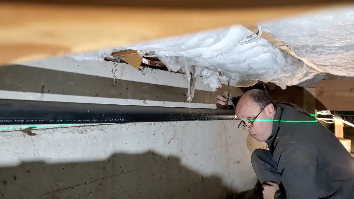 Man kneeling at the edge of a crawlspace and looking along a pipe run beneath the floor