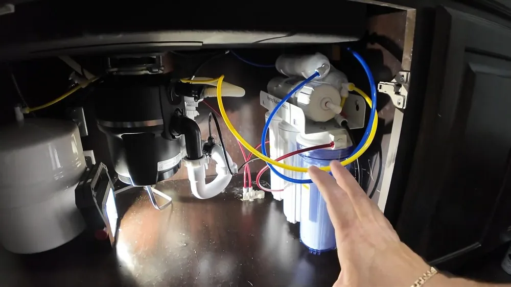 Garbage disposal and loose wiring inside a dark sink cabinet beside filter tubing