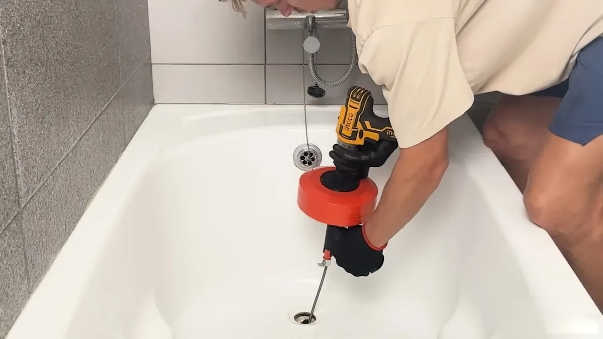 Man feeding a drill-driven drain cable into a white bathtub drain