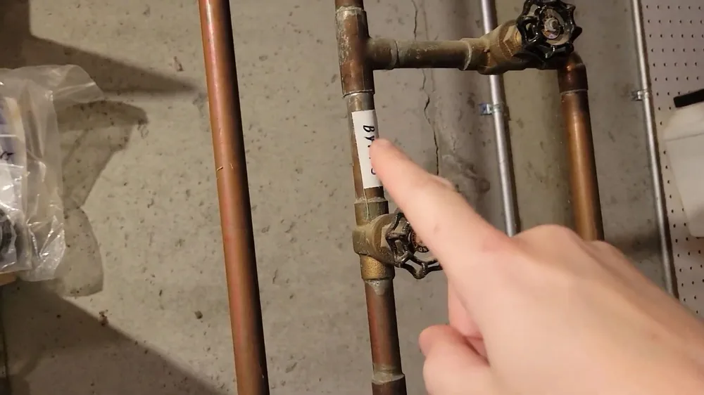 Finger pointing at two brass shutoff valves on vertical copper pipes beside a basement wall