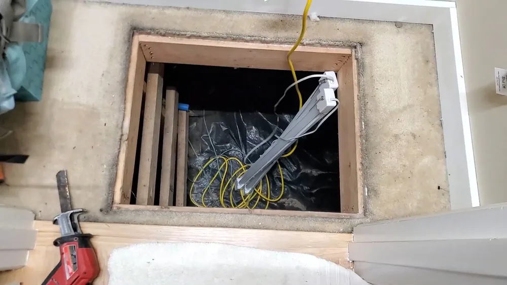 Open square floor hatch with a work light and cord leading down into a dark crawlspace
