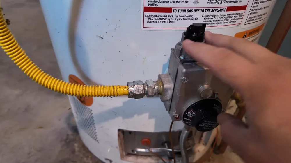 Fingers on the gas control knob of a gas-fired tank water heater beside a yellow flexible gas line