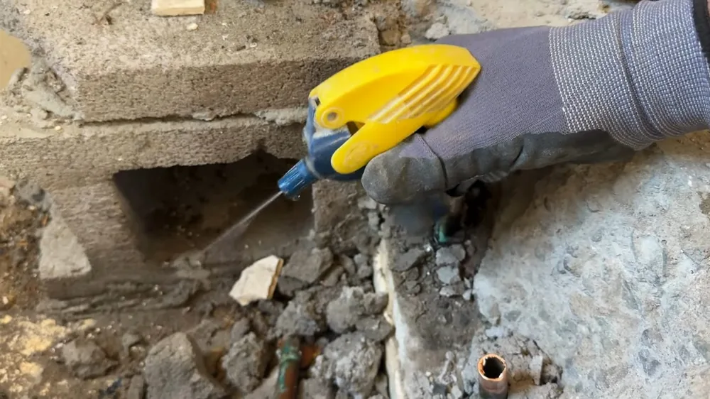 Gloved hand with a yellow chisel tool chipping a concrete slab beside a block wall to expose a pipe