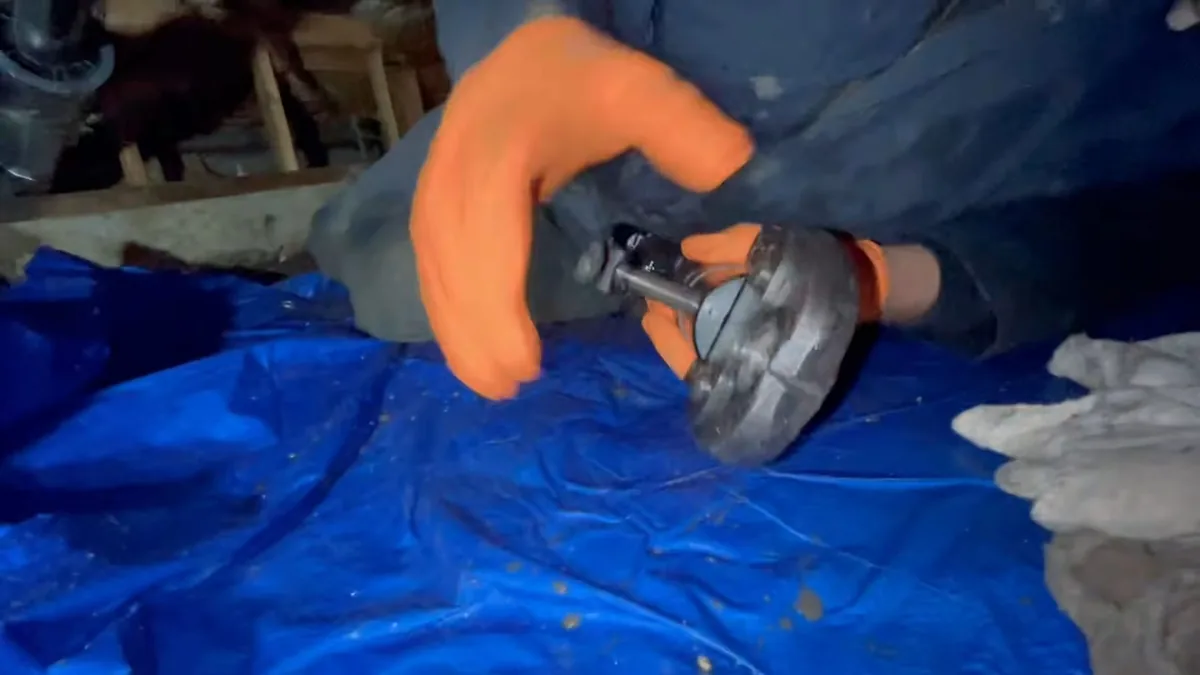 Orange-gloved hands working on a metal part over a blue tarp in a crawlspace
