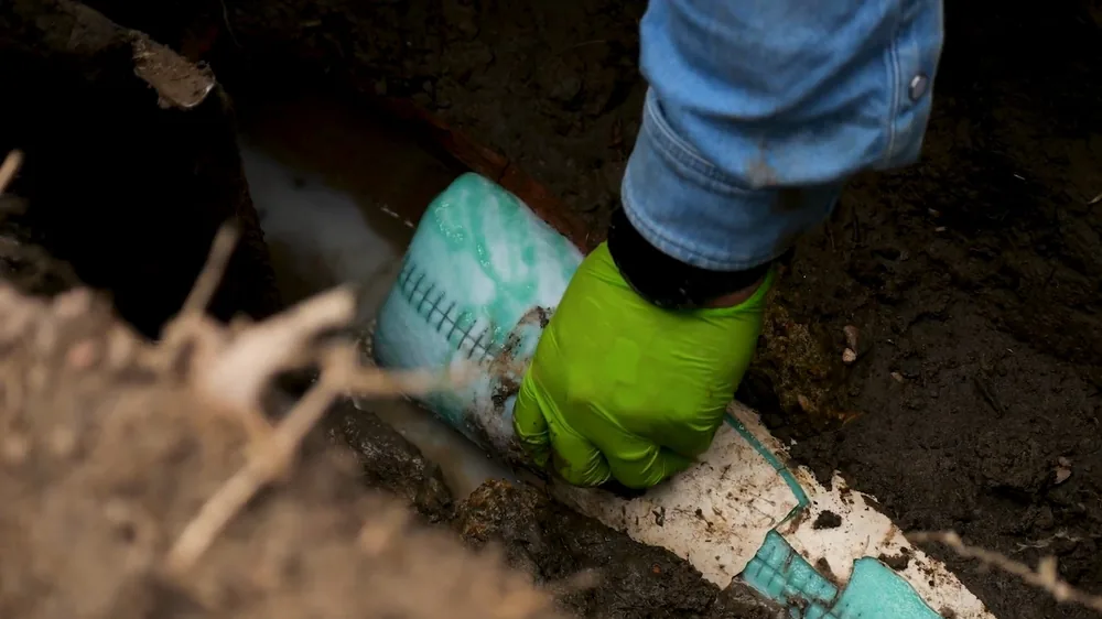 Green-gloved hand placing a pipe liner in a narrow trench beside a white pipe