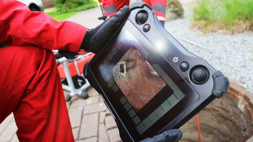 Gloved hands holding a handheld screen that shows the inside of a pipe