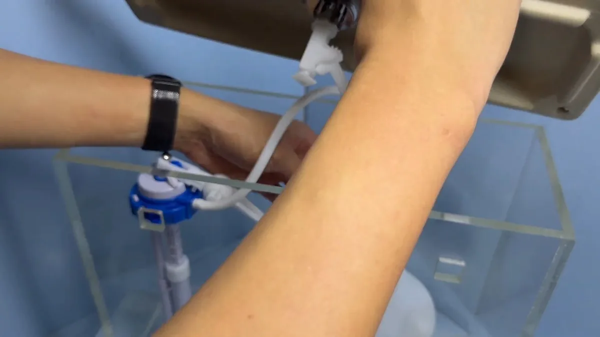Hands adjusting a fill valve inside a clear demonstration toilet tank