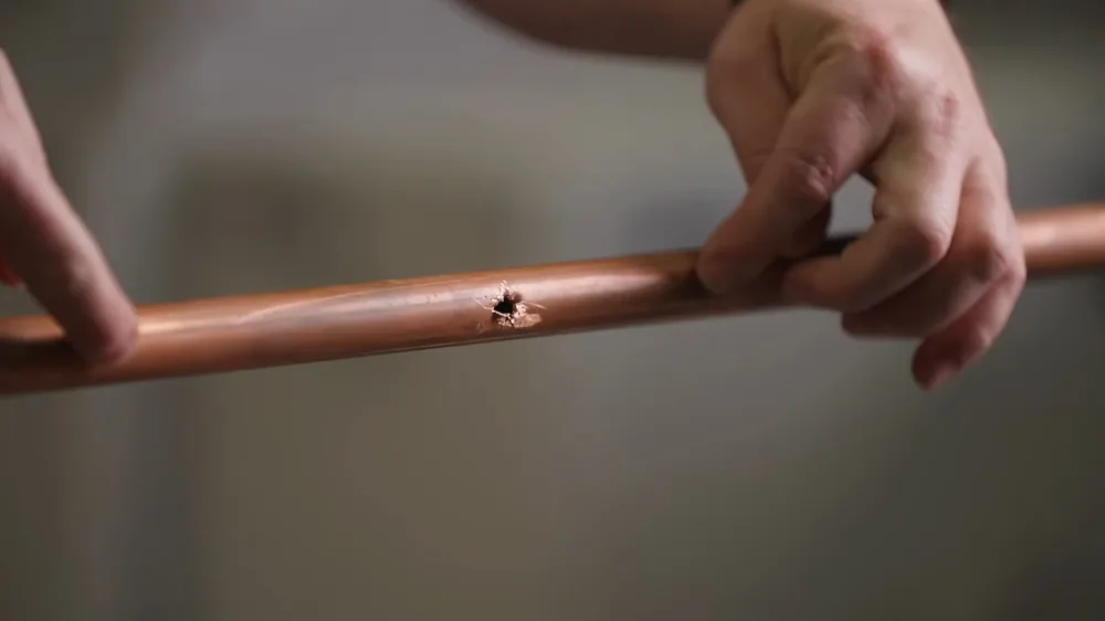 Two hands holding a length of copper pipe with a small pinhole in its side