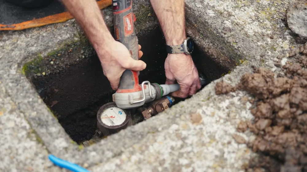 Two hands fitting a tool onto a water meter inside a concrete ground box set in the soil
