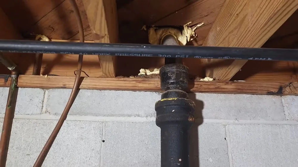 Black cast iron drain stack and copper supply tubing running beneath wood joists above a block wall