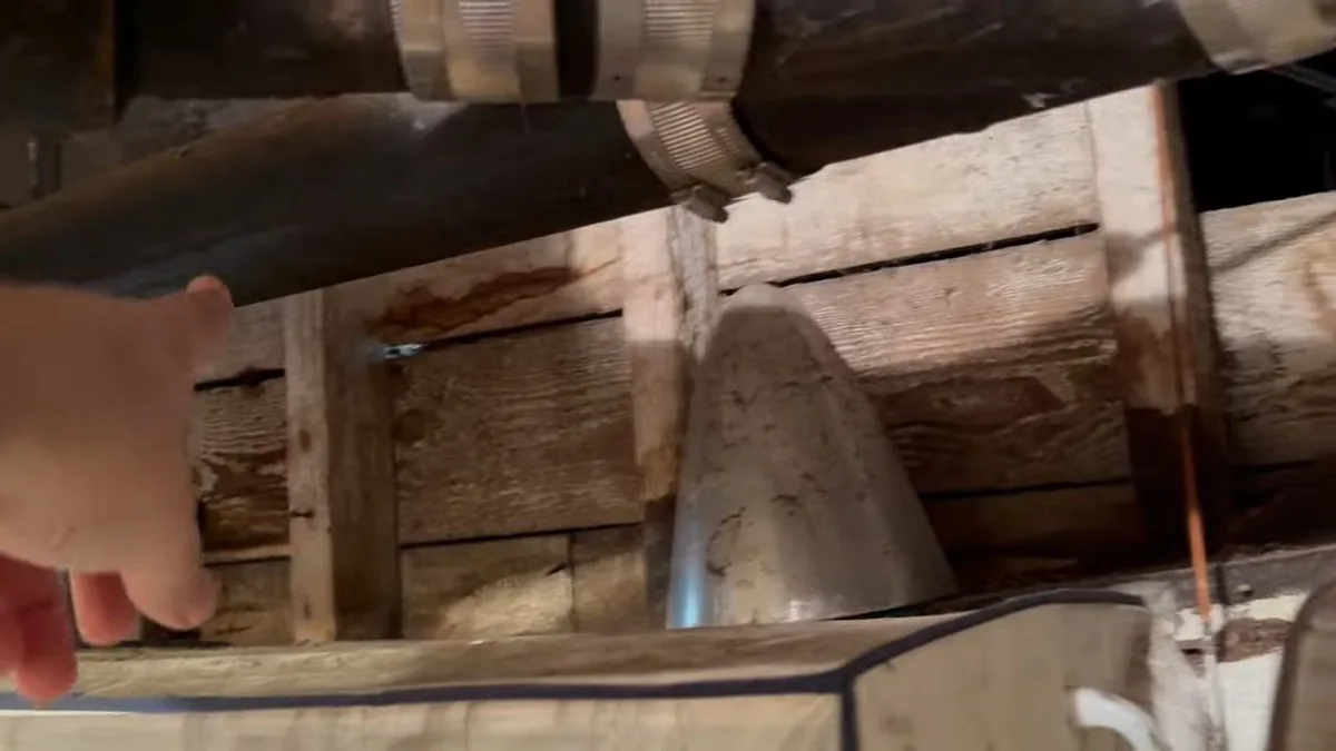 Black drain pipe with a clamped coupling running beneath old wood floor framing, with a hand reaching in at the left