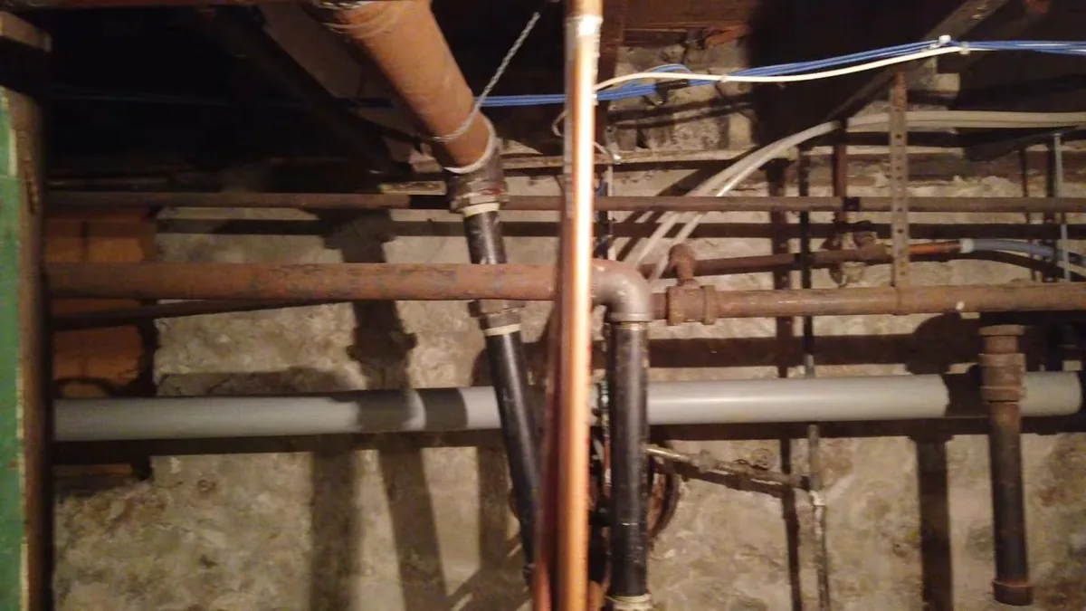 Old galvanized and copper pipes exposed beneath a basement floor between wood joists