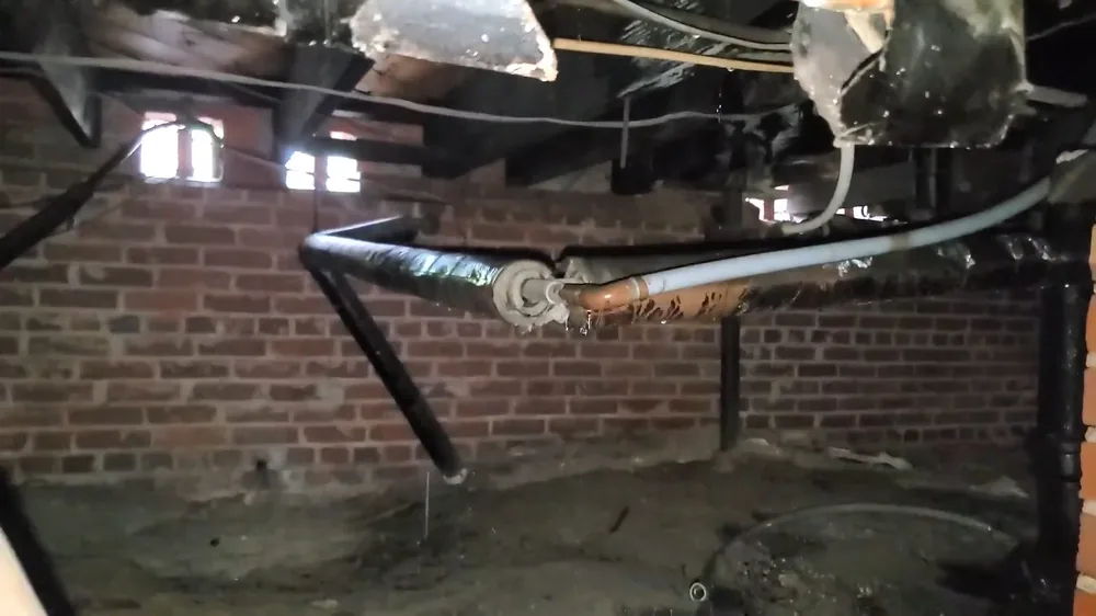 Black drain pipes and bundled supply lines hanging under floor joists above a brick crawlspace wall