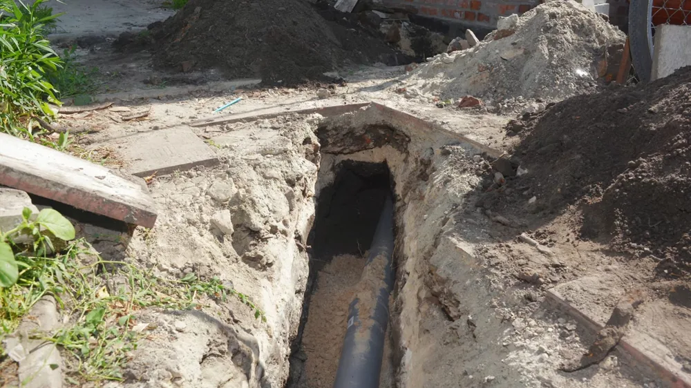 Open trench with a dark buried pipe exposed beside a pile of excavated sand and soil