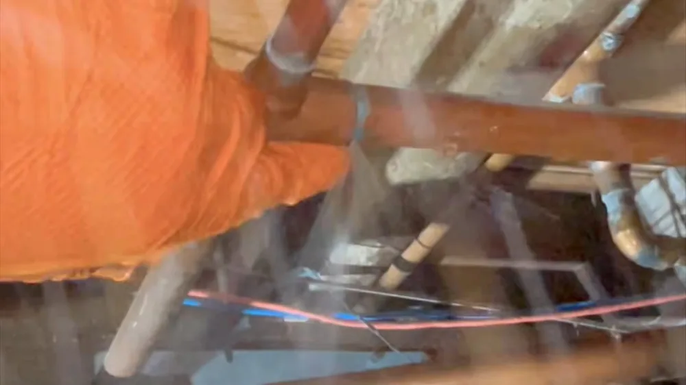Orange gloved hand held at a copper pipe spraying water beneath floor joists