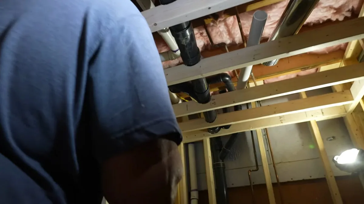 Plumber in a blue shirt looking up at pipes in an open basement ceiling with pink insulation