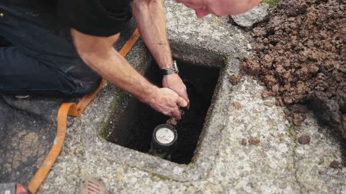 Man in dark clothes kneeling at a concrete ground box and reaching in to a water meter