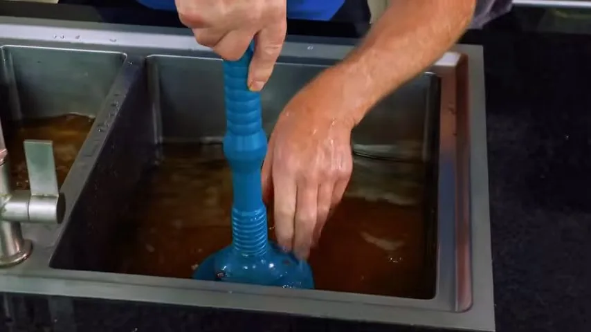 Hand working a blue plunger in a stainless kitchen sink full of brown water