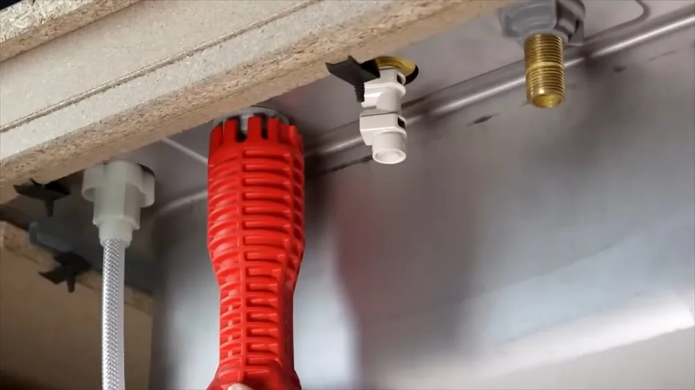 Red basin wrench held beneath a sink beside white supply fittings and a brass connector