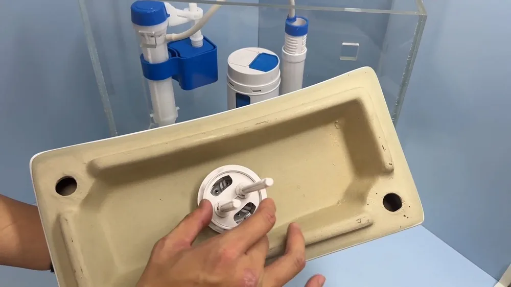 Hands fitting a replacement part into a ceramic toilet tank lid beside a clear tank model