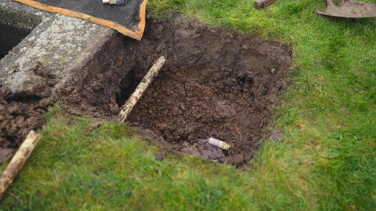 Square hole dug in a green lawn beside a concrete pad, with a short length of pipe at the bottom and a wooden stake lying across it