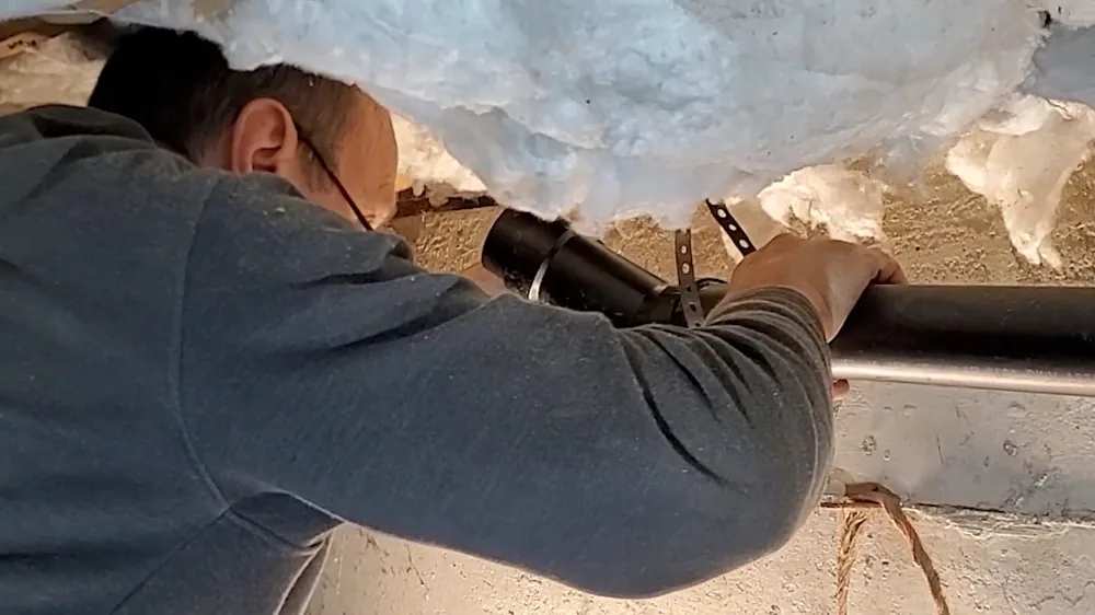 Man in a gray sweater reaching along a black drain line under insulation in a crawlspace