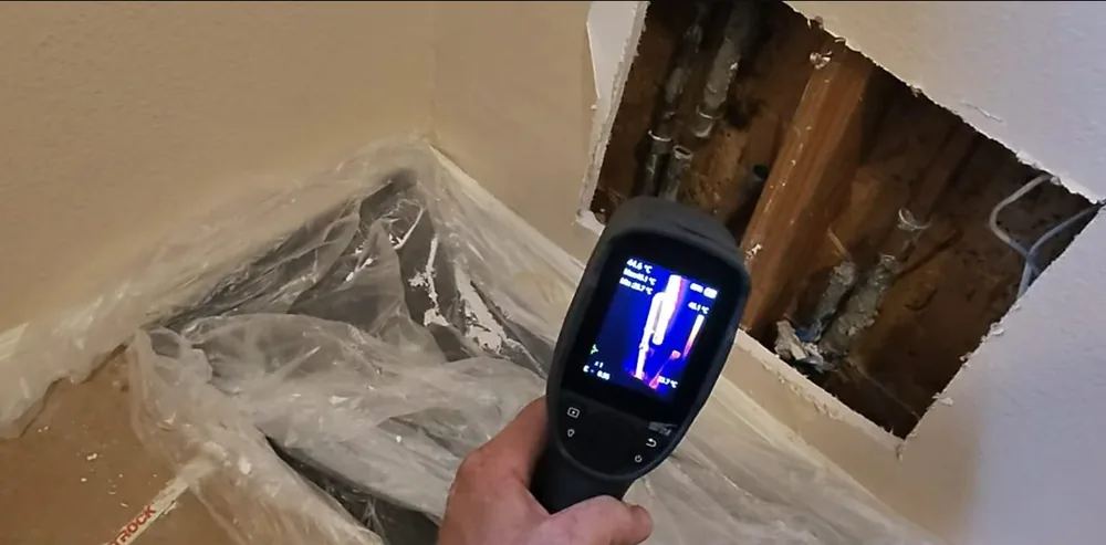 Hand holding a thermal camera toward an opened wall section above plastic sheeting