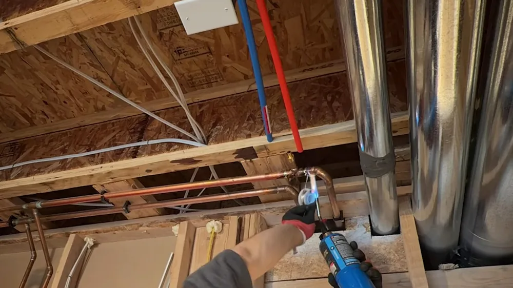 Gloved hand holding a blue torch to copper pipe between wood joists in a basement ceiling with red and blue lines overhead