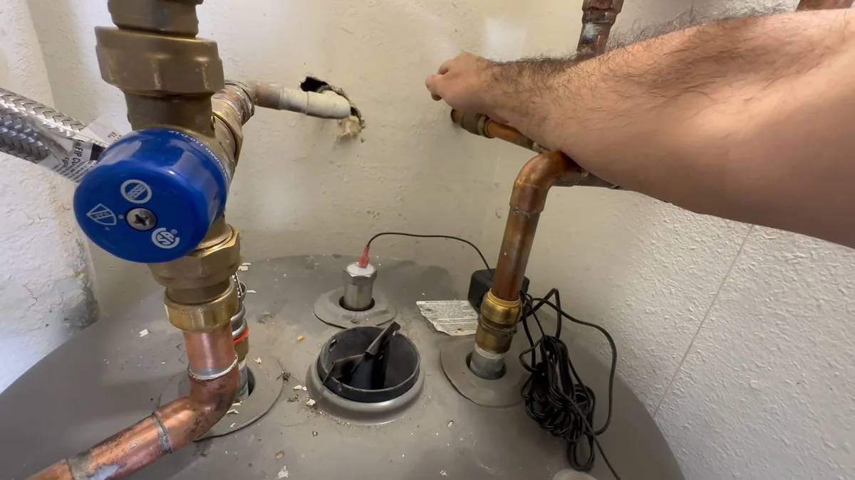 Hand reaching along the copper supply and relief fittings on top of a tank water heater