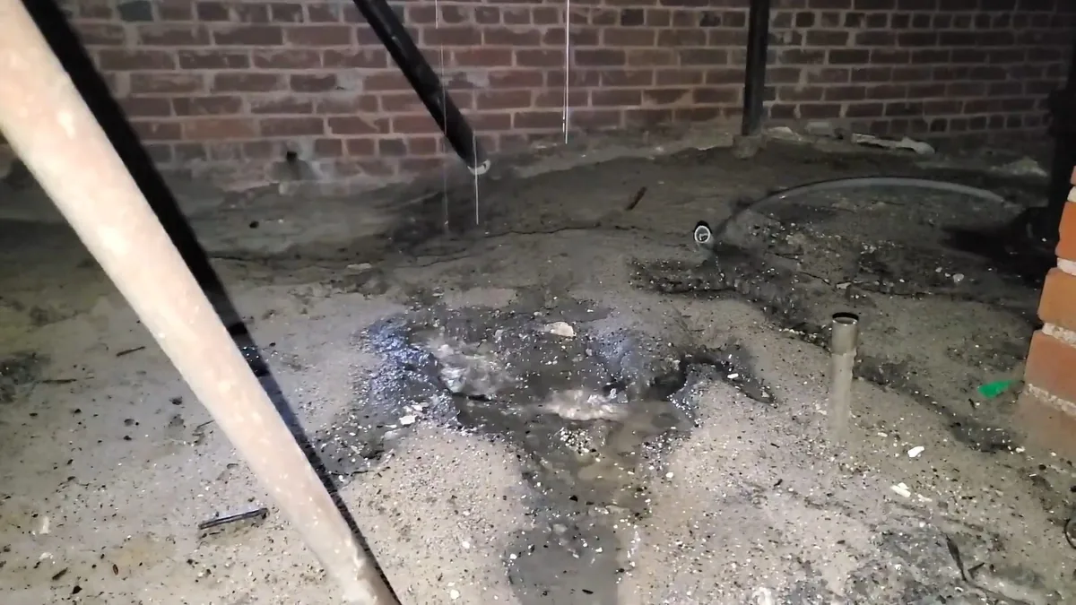 Wet dirt and pooled water under a crawlspace pipe beside a brick wall