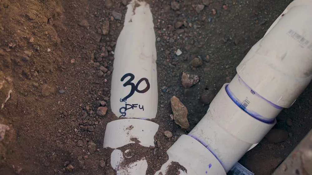 White PVC drain pipe with a handwritten number lying in dirt beside a second pipe with blue primer at the joints