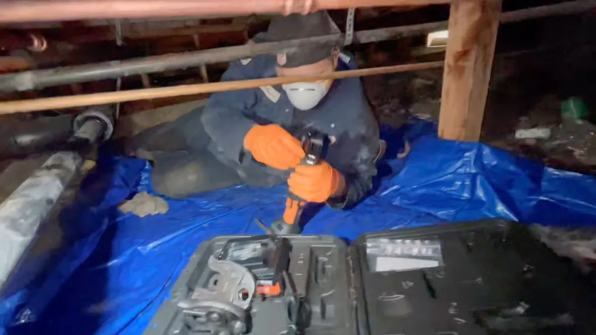 Worker in a dust mask and orange gloves repairing a pipe in a crawlspace over a blue tarp